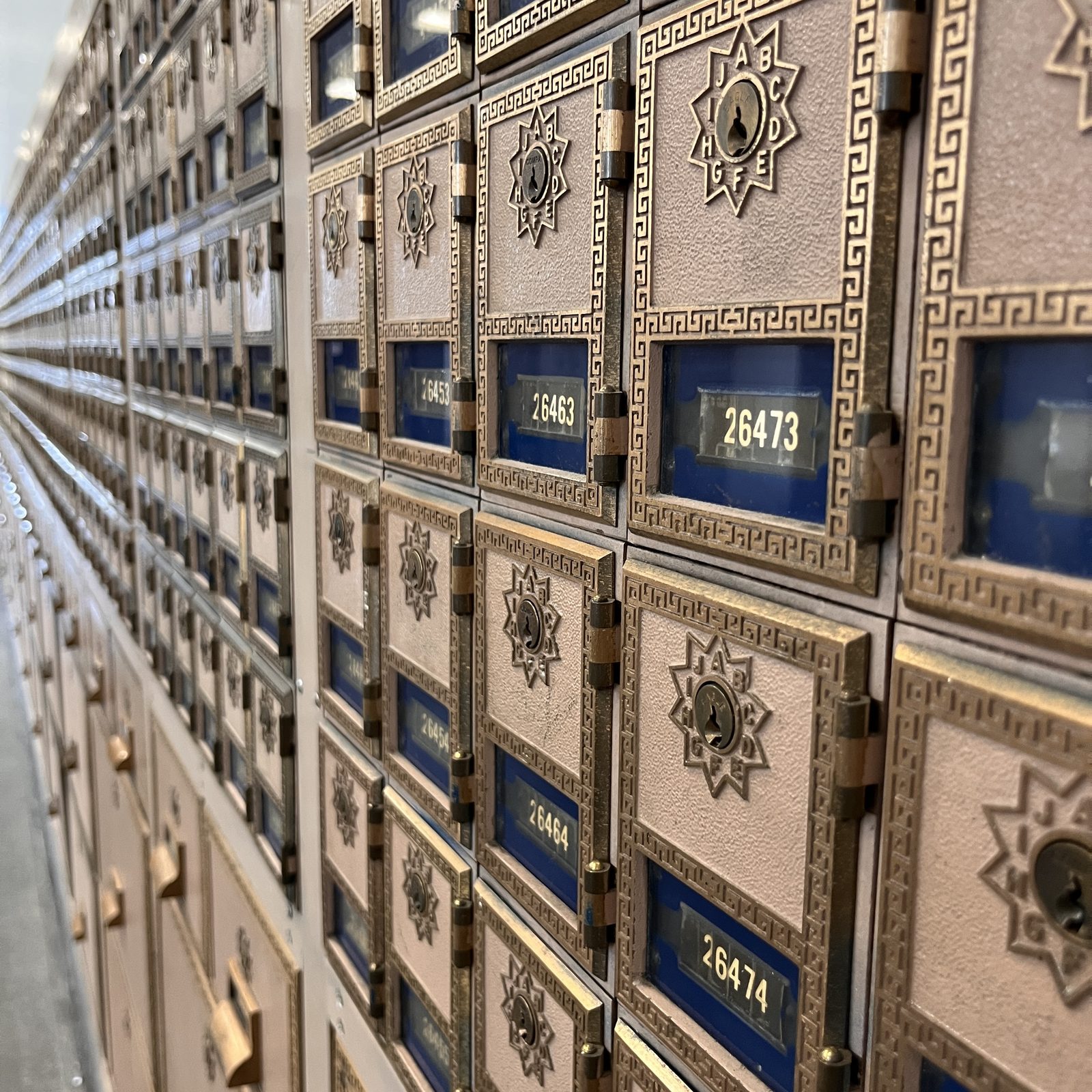 A corridor of antique brass post-office boxes receding to the vanishing point — starburst combination dials, Greek-key borders, numbered plates behind cobalt glass.
