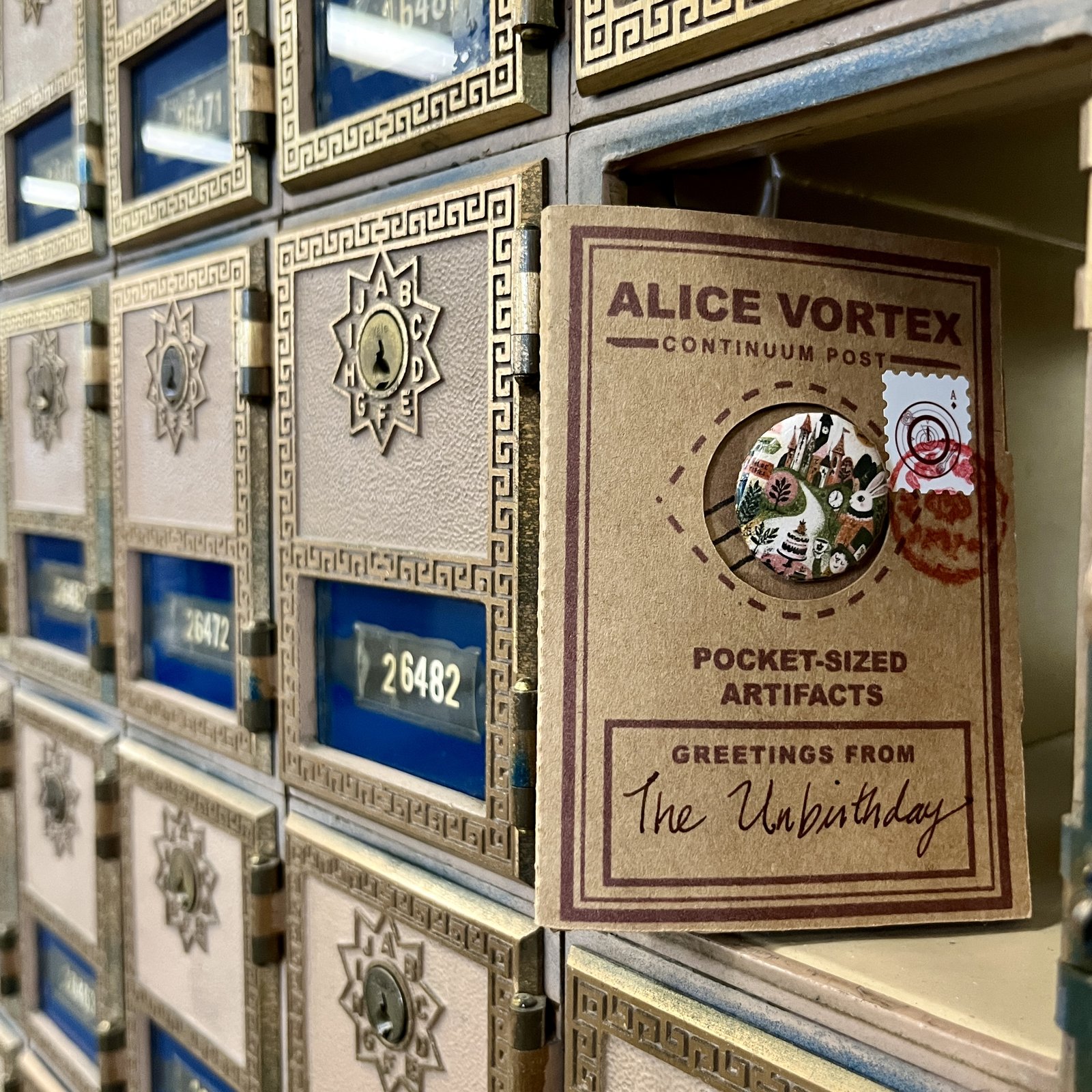 A kraft Alice Vortex Pocket-Sized Artifacts card, greetings from The Unbirthday, standing in an open antique brass post-office box amid a wall of deco brass doors