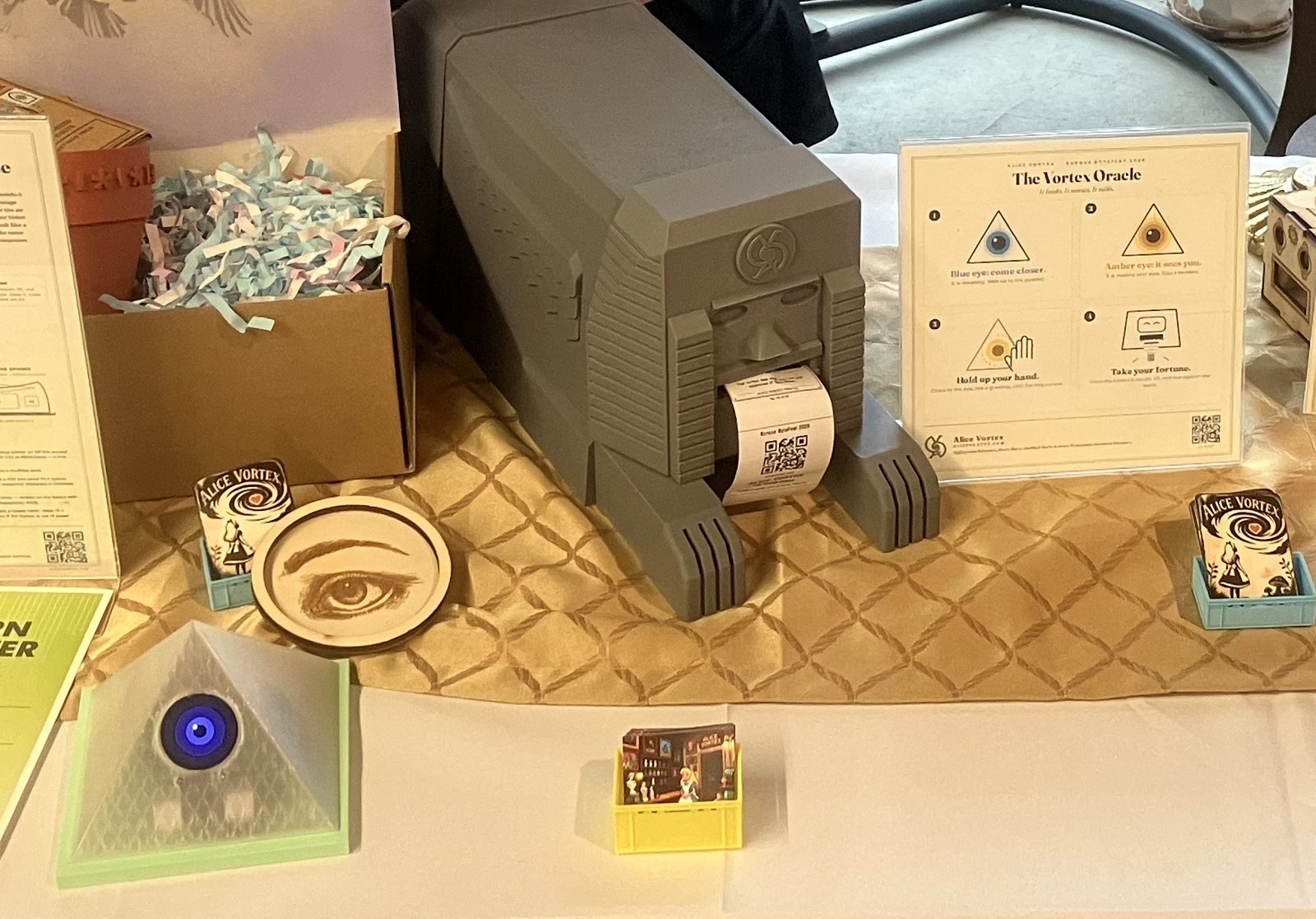 The sphinx at the Burque ByteFest table — a grey deco figure sitting very still, a fresh fortune ticket held out of its mouth, the pyramid's eye glowing blue nearby